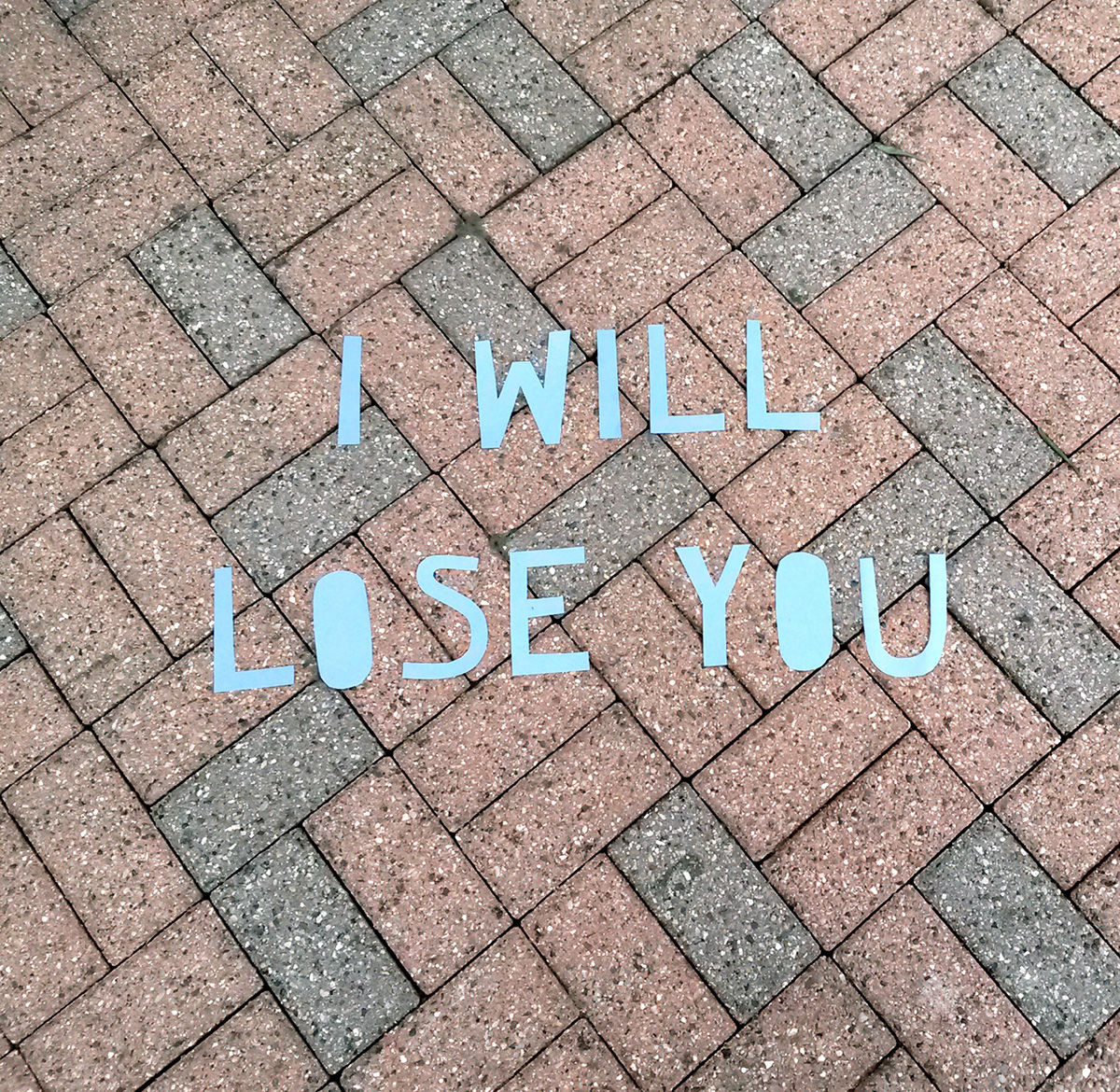 Photo of neat pink and grey brick paving, with words cut out from blue paper laid on top reading 'I WILL LOSE YOU'