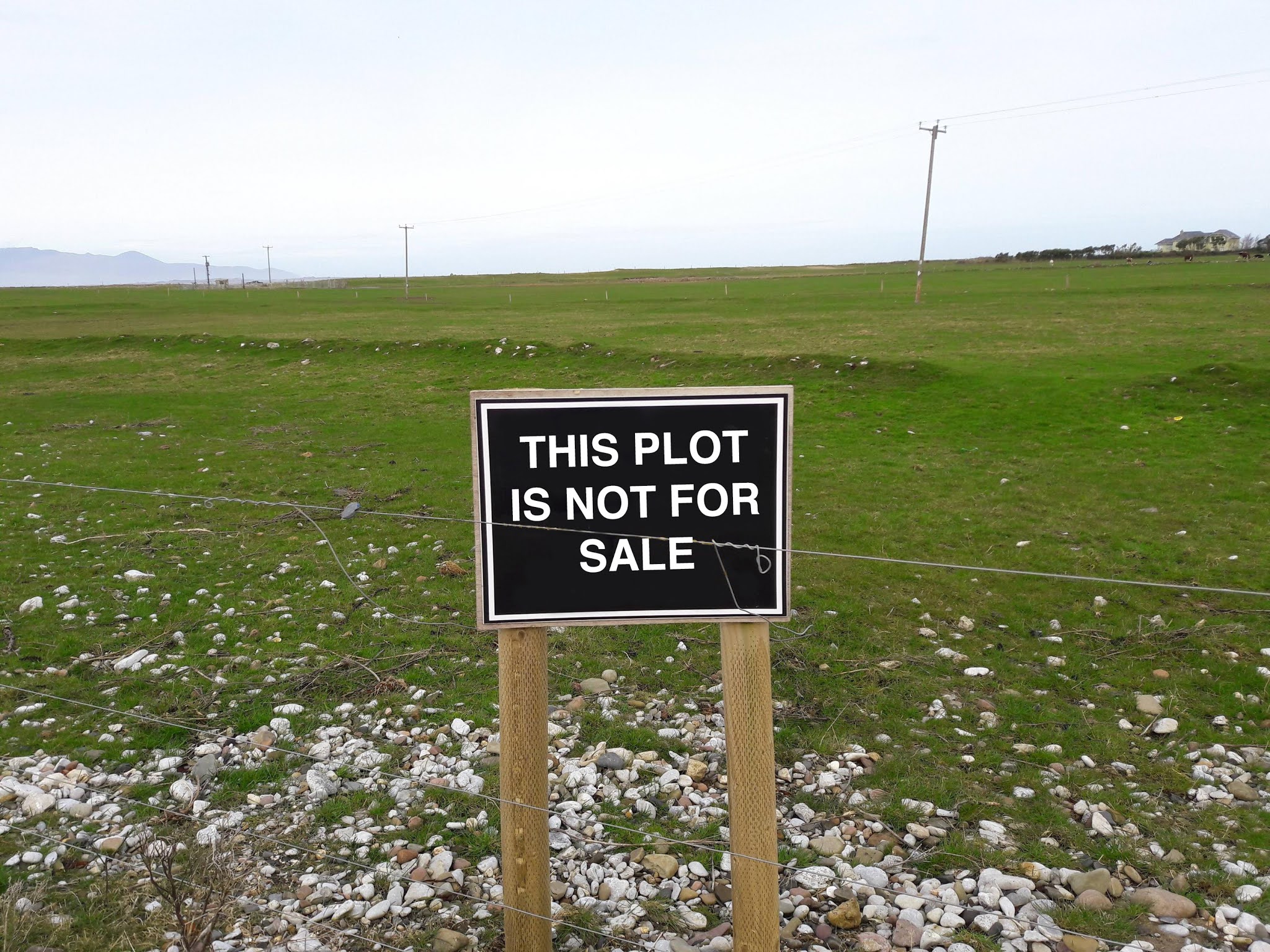 Photo of a large stretch of grass, empty except for some pylons. Some hills are just visible in the background. In the foreground some stones cover the grass and a sign reads 'THIS PLOT IS NOT FOR SALE'