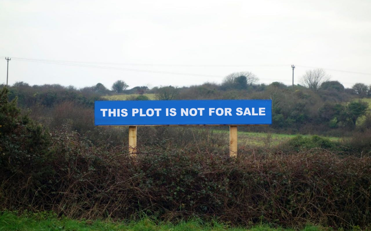 Photo of some fields and patched of dense hedges, bushes and trees, with grey skies and pylons in the background. A blue sign in the foreground reads 'THIS PLOT IS NOT FOR SALE'