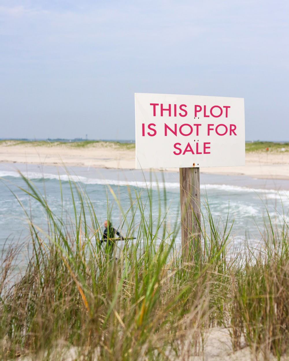 Photo of a chalky British beach, looking out from a grassy sand bank across waves to more beach and land. There is a man standing in the sea in the background and the sky is blue. In the foreground is a red and white sign saying 'THIS PLOT IS NOT FOR SALE'