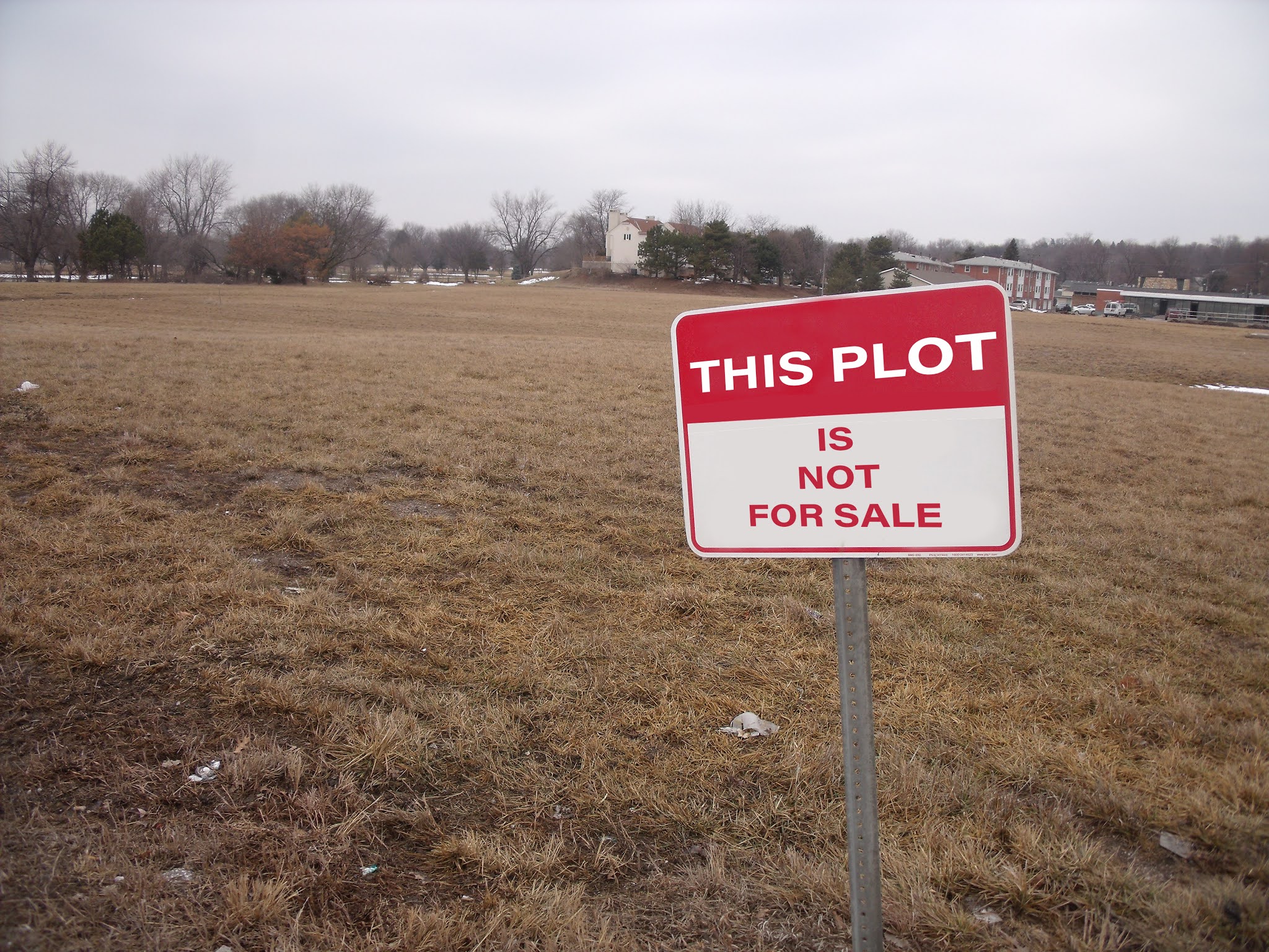 Photo of a large scrubby patch of brownish grass under grey sky with some houses and trees in the background and a red and white sign in the foreground saying 'THIS PLOT IS NOT FOR SALE'