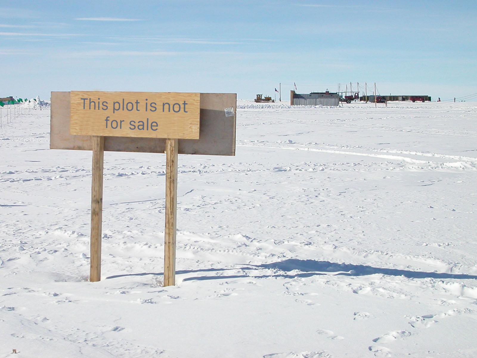 Photo of a flat, barren, snow-covered landscape with some industrial buildings and vehicles in the distance. A wooden sign in the foreground reads 'This plot is not for sale'