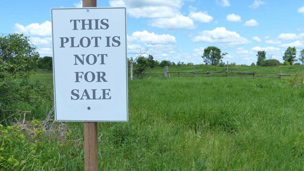 Photo of a lush green meadow with some trees, blue skies with a few clouds. A white sign very close to the camera reads 'THIS PLOT IS NOT FOR SALE'